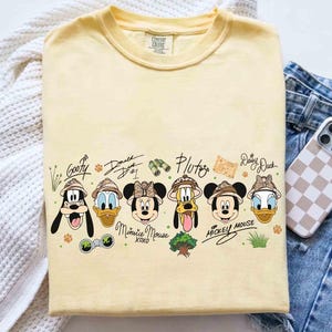 May include: A light yellow t-shirt featuring cartoon characters Goofy, Donald Duck, Minnie Mouse, Pluto, Mickey Mouse, and Daisy Duck, all wearing safari hats. The shirt includes the characters' signatures and other safari-themed graphics.