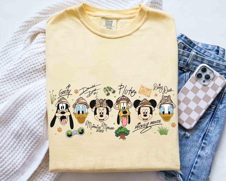 May include: A light yellow t-shirt featuring cartoon characters Goofy, Donald Duck, Minnie Mouse, Pluto, Mickey Mouse, and Daisy Duck, all wearing safari hats. The shirt includes the characters' signatures and other safari-themed graphics.