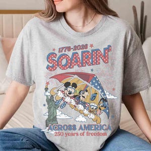 May include: A gray t-shirt featuring a graphic of Mickey Mouse, Goofy, and Donald Duck hang gliding over the Statue of Liberty. The shirt has the text "1776-2026 SOARIN' ACROSS AMERICA 250 years of freedom."