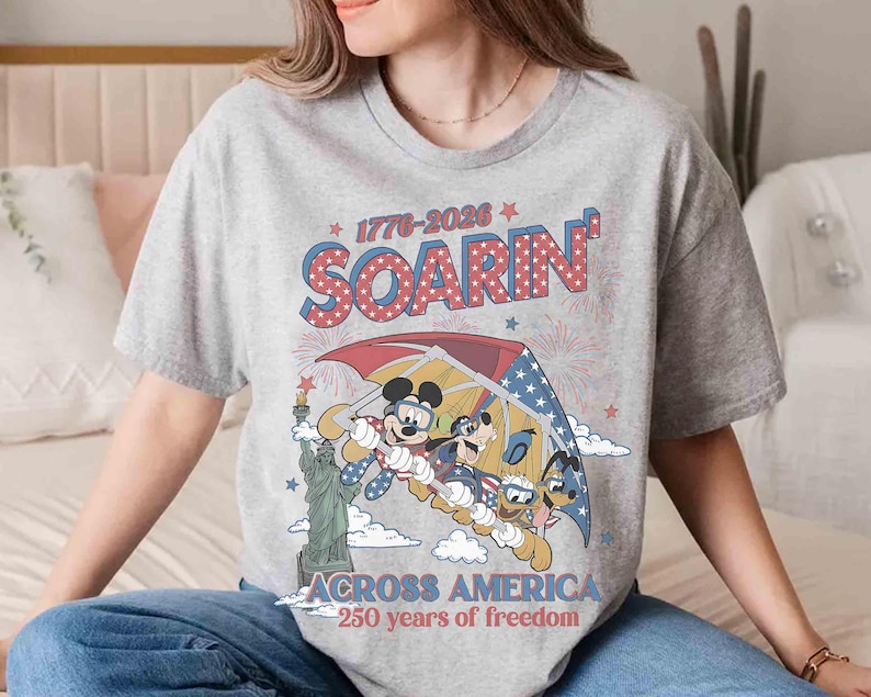 May include: A gray t-shirt featuring a graphic of Mickey Mouse, Goofy, and Donald Duck hang gliding over the Statue of Liberty. The shirt has the text "1776-2026 SOARIN' ACROSS AMERICA 250 years of freedom."