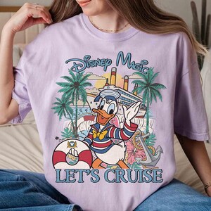 May include: A lilac-colored t-shirt featuring a graphic of Donald Duck in a sailor outfit, waving in front of a cruise ship, palm trees, and the text "Disney Magic Let's Cruise". A life preserver and anchor are also visible.