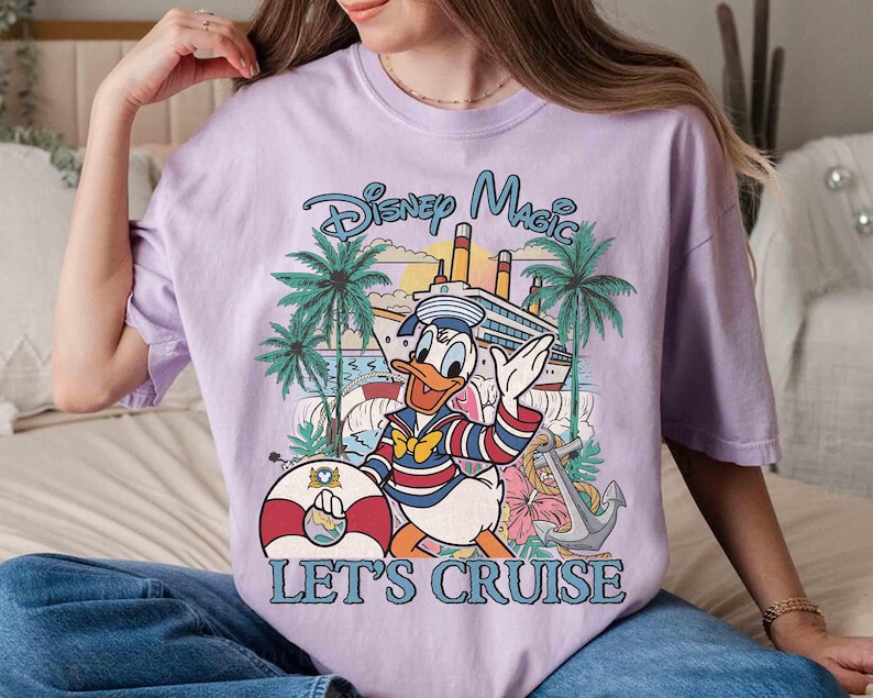 May include: A lilac-colored t-shirt featuring a graphic of Donald Duck in a sailor outfit, waving in front of a cruise ship, palm trees, and the text "Disney Magic Let's Cruise". A life preserver and anchor are also visible.