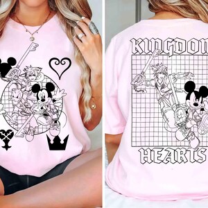 Puede incluir: Camiseta rosa claro con un gr&aacute;fico en blanco y negro de Mickey Mouse, Donald Duck y Sora de Kingdom Hearts. El dise&ntilde;o frontal incluye una llave, un coraz&oacute;n y una corona. La parte trasera de la camiseta muestra las palabras "Kingdom Hearts" con una ilustraci&oacute;n de personaje.