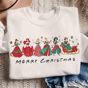 May include: White sweatshirt featuring a graphic of several princesses in Christmas attire. The princesses are in red and green dresses, with the text "MERRY CHRISTMAS" in black letters across the bottom.