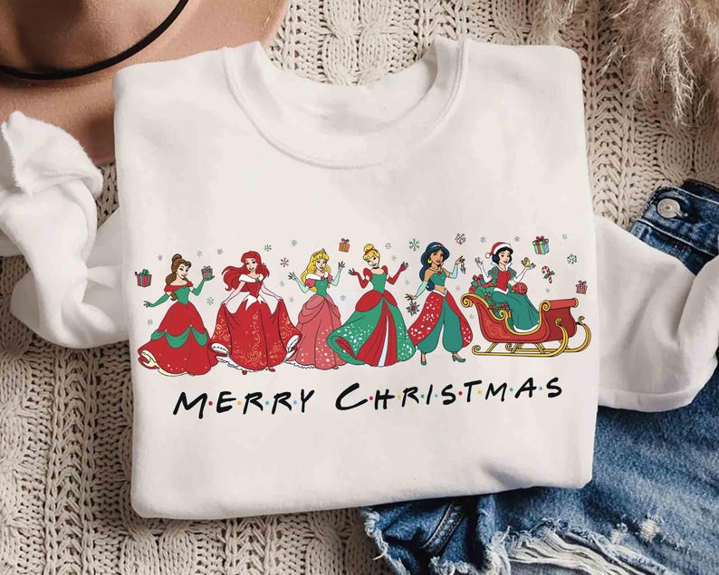 May include: White sweatshirt featuring a graphic of several princesses in Christmas attire. The princesses are in red and green dresses, with the text "MERRY CHRISTMAS" in black letters across the bottom.
