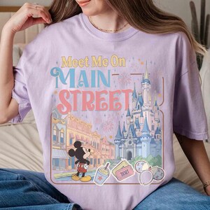 May include: A light purple t-shirt with the text "Meet Me On Main Street" above a graphic of a cartoon castle, buildings, and Mickey Mouse. The design includes fireworks and other cartoon elements.