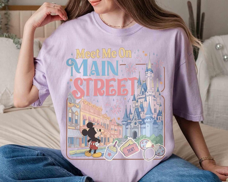 May include: A light purple t-shirt with the text "Meet Me On Main Street" above a graphic of a cartoon castle, buildings, and Mickey Mouse. The design includes fireworks and other cartoon elements.