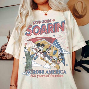 May include: Cream-colored t-shirt featuring a graphic of Mickey Mouse, Goofy, and Donald Duck soaring under a red, white, and blue parachute. The shirt includes the text "SOARIN'" and "ACROSS AMERICA - 250 years of freedom."