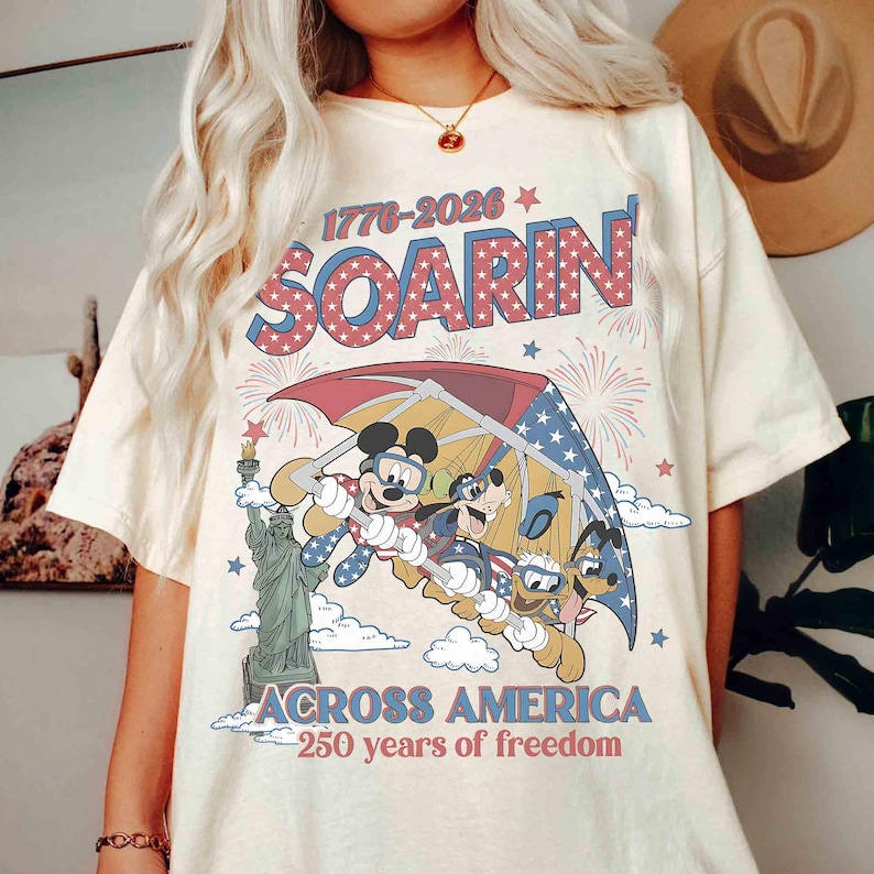 May include: Cream-colored t-shirt featuring a graphic of Mickey Mouse, Goofy, and Donald Duck soaring under a red, white, and blue parachute. The shirt includes the text "SOARIN'" and "ACROSS AMERICA - 250 years of freedom."