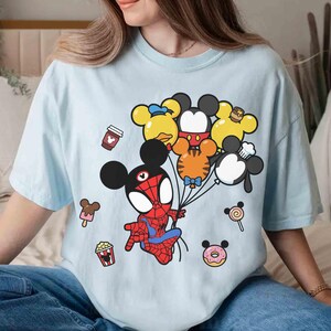 Puede incluir: Camiseta azul claro con un personaje de dibujos animados de Spider-Man sosteniendo globos con forma de Mickey Mouse y sus amigos. La camiseta tambi&eacute;n tiene gr&aacute;ficos de una taza de caf&eacute;, palomitas de ma&iacute;z, helado, una piruleta y una rosquilla.