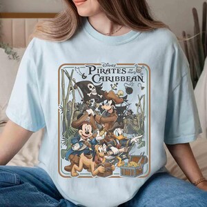May include: Light blue t-shirt featuring a graphic from Disney's Pirates of the Caribbean. The design includes Mickey Mouse, Goofy, Donald Duck, and Pluto dressed as pirates, with a treasure chest and pirate ship. The text "Disney Pirates of the Caribbean" is at the top.