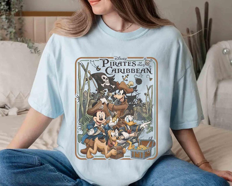 May include: Light blue t-shirt featuring a graphic from Disney's Pirates of the Caribbean. The design includes Mickey Mouse, Goofy, Donald Duck, and Pluto dressed as pirates, with a treasure chest and pirate ship. The text "Disney Pirates of the Caribbean" is at the top.