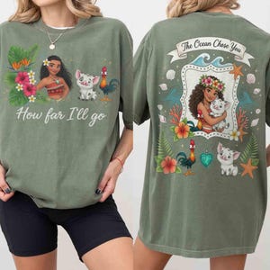Op de afbeelding: Olijfgroen T-shirt met een afbeelding van een Polynesische vrouw, een varken en een haan, met de tekst "How far I'll go" op de voorkant. De achterkant heeft een vergelijkbaar ontwerp met de tekst "The Ocean Chose You".