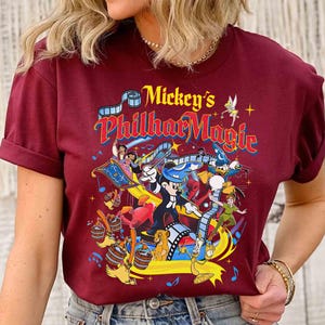 K&ouml;nnte beinhalten: Burgunderfarbenes T-Shirt mit einer farbenfrohen Grafik von "Mickey's PhilharMagic". Das Design zeigt Micky Maus, Peter Pan, Donald Duck und andere Disney-Figuren. Der Text "Mickey's PhilharMagic" ist in einer lebendigen, mehrfarbigen Schriftart gehalten.