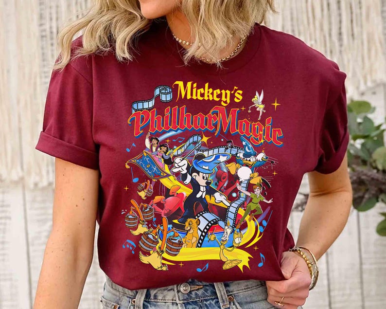 K&ouml;nnte beinhalten: Burgunderfarbenes T-Shirt mit einer farbenfrohen Grafik von "Mickey's PhilharMagic". Das Design zeigt Micky Maus, Peter Pan, Donald Duck und andere Disney-Figuren. Der Text "Mickey's PhilharMagic" ist in einer lebendigen, mehrfarbigen Schriftart gehalten.