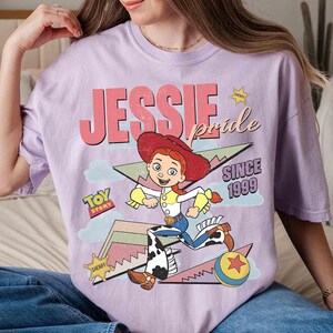 May include: A lilac-colored t-shirt featuring a graphic of the character Jessie from Toy Story. The design includes the text "JESSIE pride SINCE 1999" and a cartoon image of Jessie in her cowgirl outfit. A "Toy Story" logo is also present.
