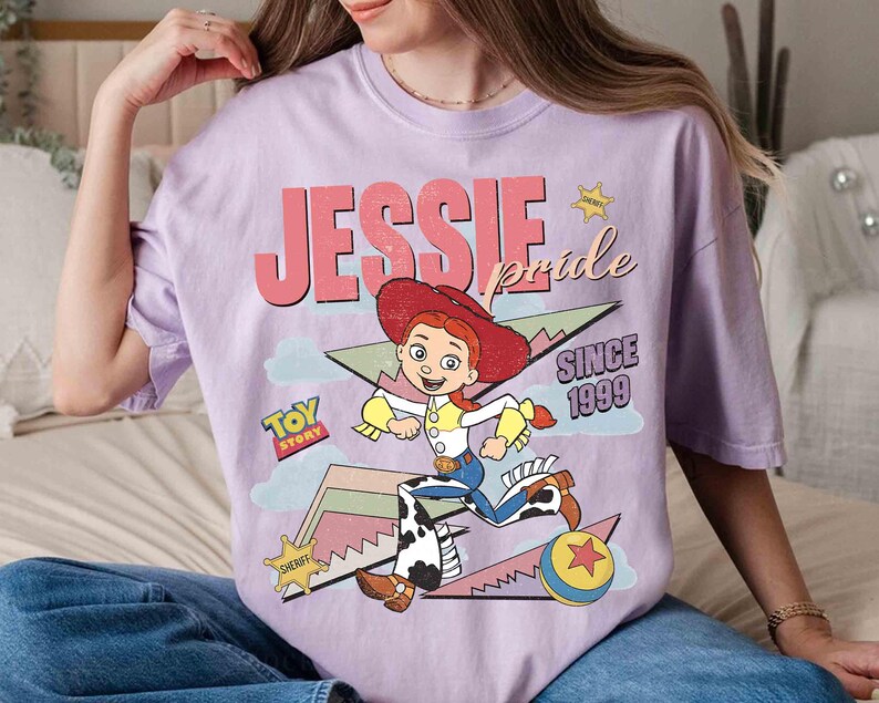 May include: A lilac-colored t-shirt featuring a graphic of the character Jessie from Toy Story. The design includes the text "JESSIE pride SINCE 1999" and a cartoon image of Jessie in her cowgirl outfit. A "Toy Story" logo is also present.