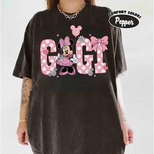 May include: Black t-shirt featuring a Minnie Mouse graphic with the word "GIGI" in pink polka dot letters. The design includes a bow, flowers, and a balloon. The shirt has the words "Comfort Colors" and "Pepper" in a circle.