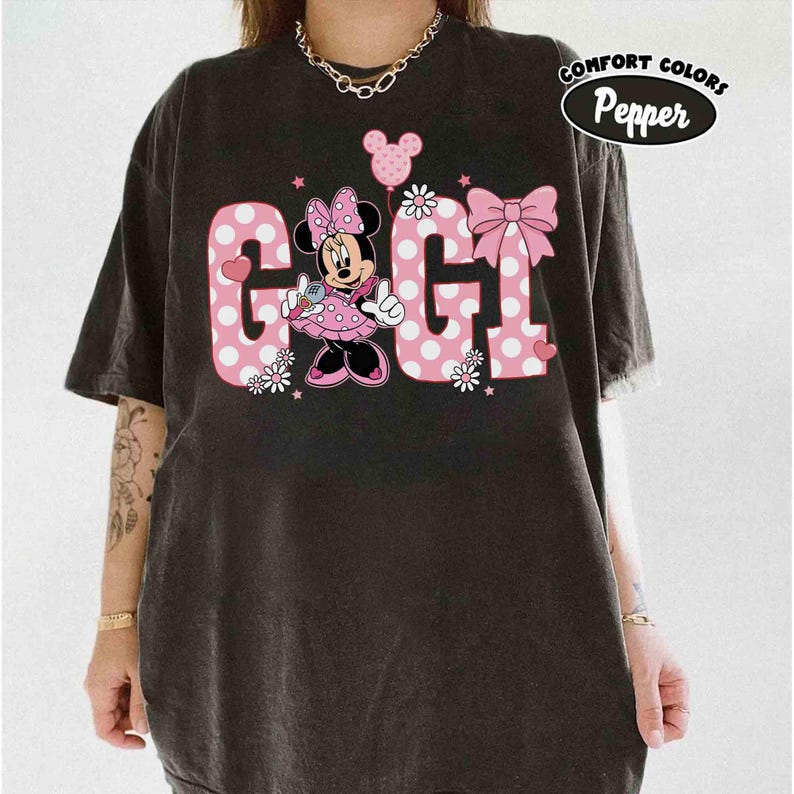 May include: Black t-shirt featuring a Minnie Mouse graphic with the word "GIGI" in pink polka dot letters. The design includes a bow, flowers, and a balloon. The shirt has the words "Comfort Colors" and "Pepper" in a circle.
