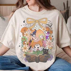 K&ouml;nnte beinhalten: Cremefarbenes T-Shirt mit einem Halloween-Motiv mit Zeichentrickfiguren, Blumen und einer schwarzen Katze. Das Design umfasst eine Schleife, Flederm&auml;use und ein Zauberbuch. Das Shirt hat eine lockere Passform.