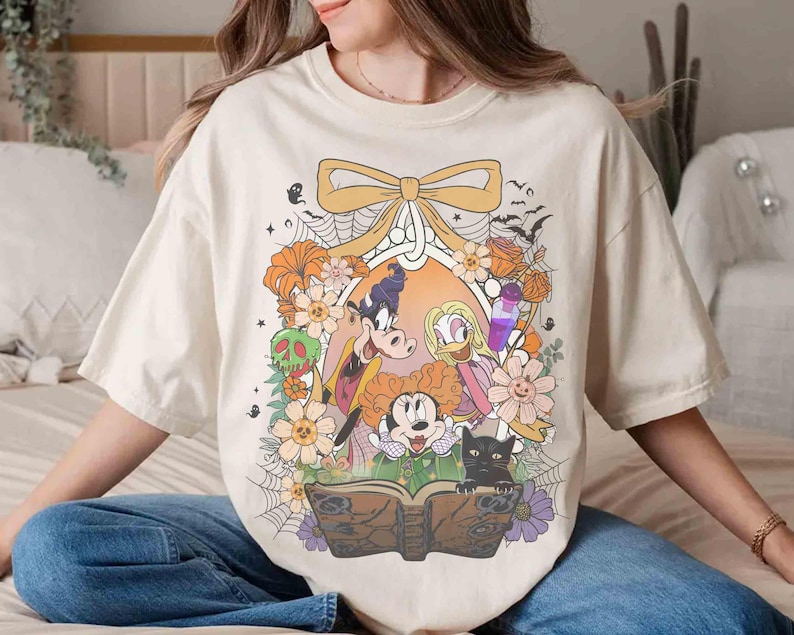 K&ouml;nnte beinhalten: Cremefarbenes T-Shirt mit einem Halloween-Motiv mit Zeichentrickfiguren, Blumen und einer schwarzen Katze. Das Design umfasst eine Schleife, Flederm&auml;use und ein Zauberbuch. Das Shirt hat eine lockere Passform.