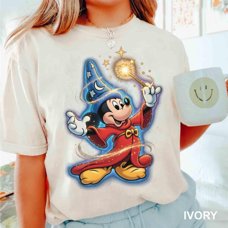 May include: Ivory-colored t-shirt featuring a cartoon Mickey Mouse dressed as a wizard, holding a glowing wand. Mickey wears a blue wizard hat with stars and a red robe. A white mug with a smiley face is visible.
