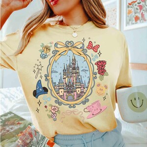 May include: A yellow t-shirt featuring a graphic print of a castle, flowers, and Mickey Mouse characters. The castle is enclosed within a gold frame adorned with a bow. The shirt has a relaxed fit and short sleeves.