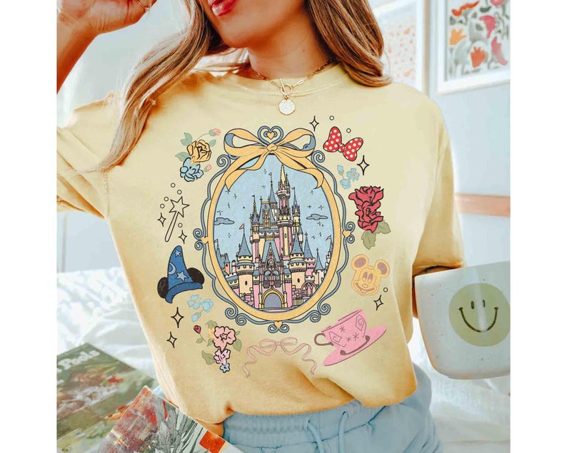May include: A yellow t-shirt featuring a graphic print of a castle, flowers, and Mickey Mouse characters. The castle is enclosed within a gold frame adorned with a bow. The shirt has a relaxed fit and short sleeves.