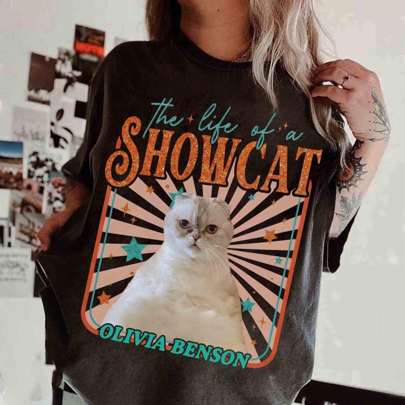 Może przedstawiać: Ciemnoszara koszulka z grafiką białego kota i napisem "the life of a SHOWCAT" w kolorze pomarańczowym i turkusowym. Grafika kota jest w stylu retro z turkusowo-pomarańczową obw&oacute;dką i napisem "OLIVIA-BENSON" poniżej.