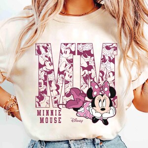 May include: Cream-colored t-shirt featuring a graphic of Minnie Mouse. The letters "MM" are filled with Minnie Mouse illustrations. Below the letters, the text reads "MINNIE MOUSE" and the Disney logo.