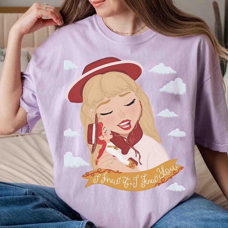 May include: A light purple t-shirt featuring a graphic illustration of a blonde person wearing a red hat and holding a small doll with a matching red hat. The design includes white clouds and a banner with the text "I Knew It, I Knew You".