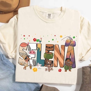 Puede incluir: Camiseta color crema con el nombre "SELENA" en letras tem&aacute;ticas y coloridas. El dise&ntilde;o incorpora personajes de dibujos animados y detalles florales. La camiseta se muestra sobre una superficie de mezclilla azul claro.