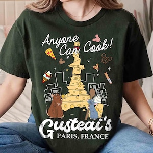 Può includere: Una maglietta verde scuro con una stampa grafica di un topo dei cartoni animati che tiene una torre di formaggio. Il testo "Anyone Can Cook! Gusteau's Paris, France" è stampato sulla maglietta.