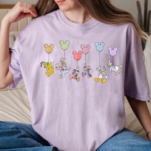 May include: A light purple t-shirt featuring a colorful graphic of Disney characters holding Mickey Mouse-shaped balloons. The characters include Pluto, Goofy, Mickey Mouse, Minnie Mouse, Donald Duck, and Daisy Duck.