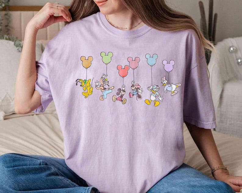 May include: A light purple t-shirt featuring a colorful graphic of Disney characters holding Mickey Mouse-shaped balloons. The characters include Pluto, Goofy, Mickey Mouse, Minnie Mouse, Donald Duck, and Daisy Duck.