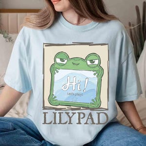 May include: Light blue t-shirt featuring a green frog holding a sign that says "Hi!" in white script. Below the frog, the text reads "Let's play!" and "LILYPAD" in brown block letters. The shirt has short sleeves.