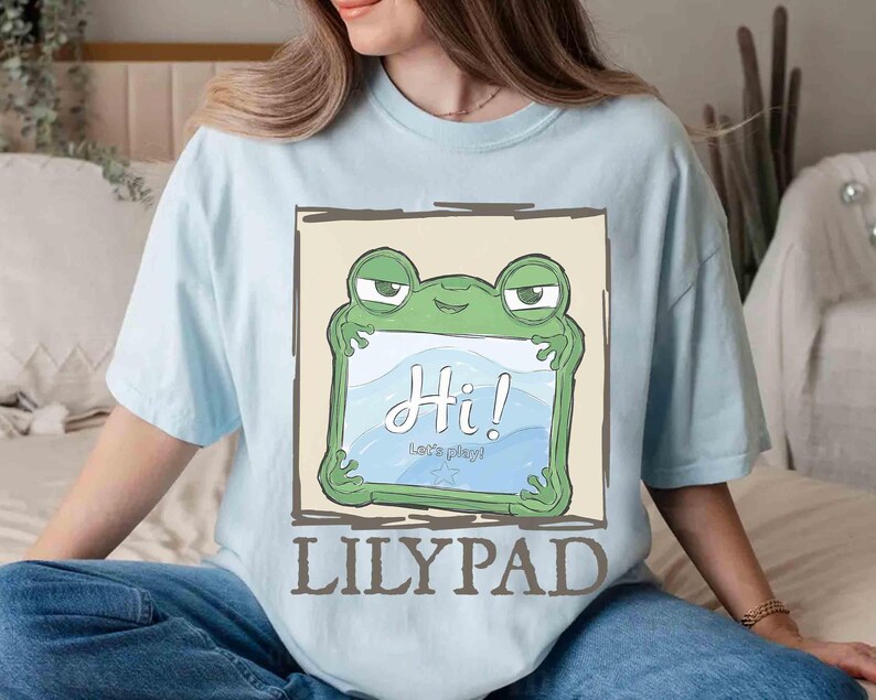 May include: Light blue t-shirt featuring a green frog holding a sign that says "Hi!" in white script. Below the frog, the text reads "Let's play!" and "LILYPAD" in brown block letters. The shirt has short sleeves.