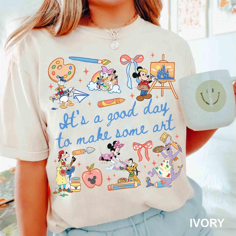 May include: Ivory-colored t-shirt featuring various Disney characters engaged in art activities. The shirt includes the phrase "It's a good day to make some art" in blue script. A white mug with a smiley face is also visible.