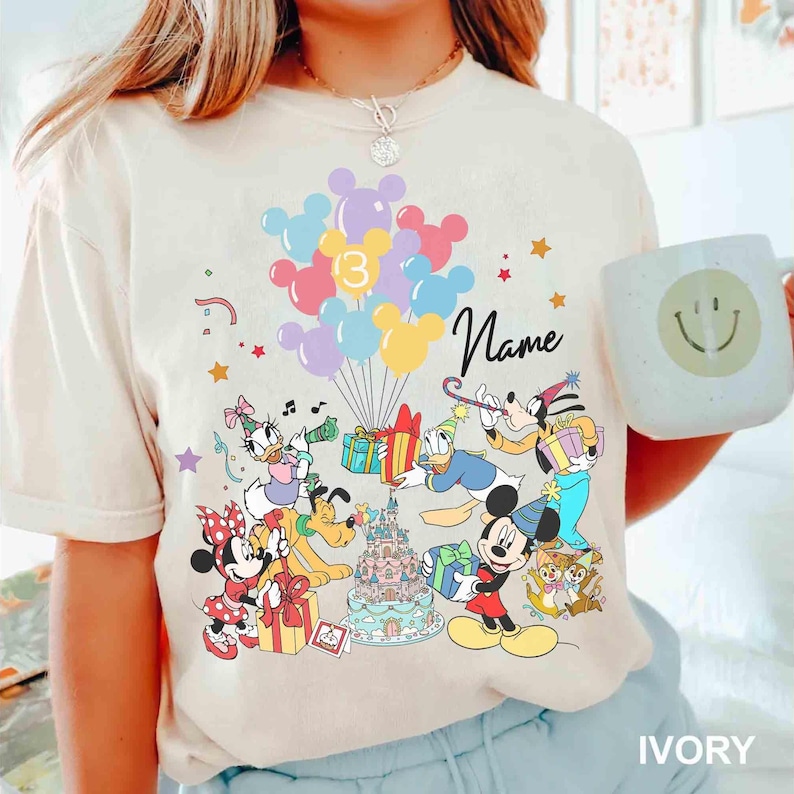 May include: Ivory-colored t-shirt featuring a colorful Disney-themed birthday design. The graphic includes Mickey Mouse, Minnie Mouse, Donald Duck, Goofy, and other characters, along with balloons, gifts, and a birthday cake. The word "Name" is written in cursive.