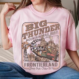 May include: A light pink t-shirt featuring a vintage-style graphic for Big Thunder Mountain Railroad in Frontierland. The design includes a train with cartoon characters, a windmill, and the text "Join Our Crew 1979."