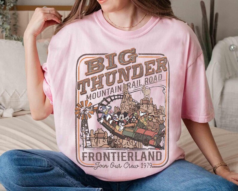 May include: A light pink t-shirt featuring a vintage-style graphic for Big Thunder Mountain Railroad in Frontierland. The design includes a train with cartoon characters, a windmill, and the text "Join Our Crew 1979."