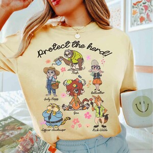 Peut inclure: T-shirt jaune avec une illustration de personnages de dessin anim&eacute; du film Zootopia. Le texte "Protect the herd!" est imprim&eacute; sur le t-shirt. Les personnages incluent Judy Hopps, Nick Wilde, Flash, Yax, Bellwether et Officer Clawhauser.