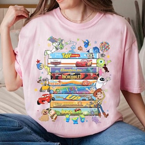 May include: A light pink t-shirt featuring a stack of colorful movie titles and characters from Disney Pixar films, including Toy Story, Monsters Inc., and Up. The design includes characters like Buzz Lightyear, Mike Wazowski, and Woody.