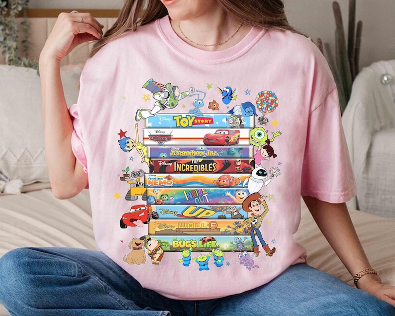 May include: A light pink t-shirt featuring a stack of colorful movie titles and characters from Disney Pixar films, including Toy Story, Monsters Inc., and Up. The design includes characters like Buzz Lightyear, Mike Wazowski, and Woody.