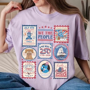 以下が含まれることがあります： 愛国的な切手デザインのコラージュが特徴の、薄紫色のTシャツ。「7月4日」や「Made in America」などのテキストとともに、自由の鐘、船、アメリカ国旗の画像が含まれています。