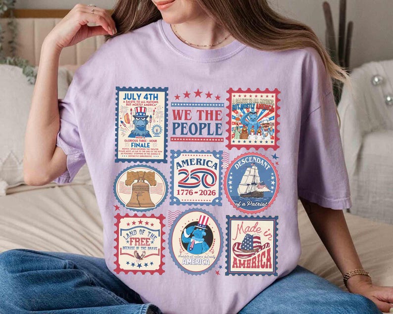 以下が含まれることがあります： 愛国的な切手デザインのコラージュが特徴の、薄紫色のTシャツ。「7月4日」や「Made in America」などのテキストとともに、自由の鐘、船、アメリカ国旗の画像が含まれています。