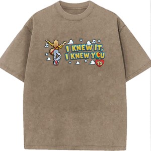 May include: A brown mineral-washed t-shirt featuring an embroidered graphic of a person with long blonde hair playing a guitar, surrounded by clouds, stars, and musical notes. The text reads I KNEW IT, I KNEW YOU TS, with a red heart icon.