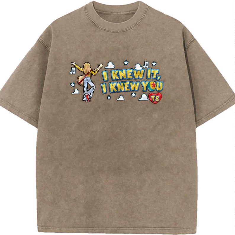 May include: A brown mineral-washed t-shirt featuring an embroidered graphic of a person with long blonde hair playing a guitar, surrounded by clouds, stars, and musical notes. The text reads I KNEW IT, I KNEW YOU TS, with a red heart icon.