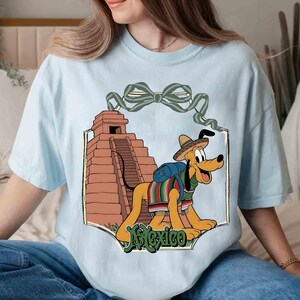 May include: Light blue t-shirt featuring a cartoon dog wearing a sombrero and a serape, with a pyramid in the background. The word "Mexico" is written in green script at the bottom of the design. A green bow is at the top of the design.