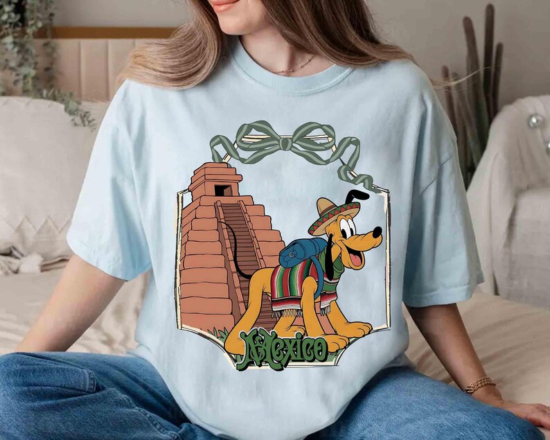 May include: Light blue t-shirt featuring a cartoon dog wearing a sombrero and a serape, with a pyramid in the background. The word "Mexico" is written in green script at the bottom of the design. A green bow is at the top of the design.